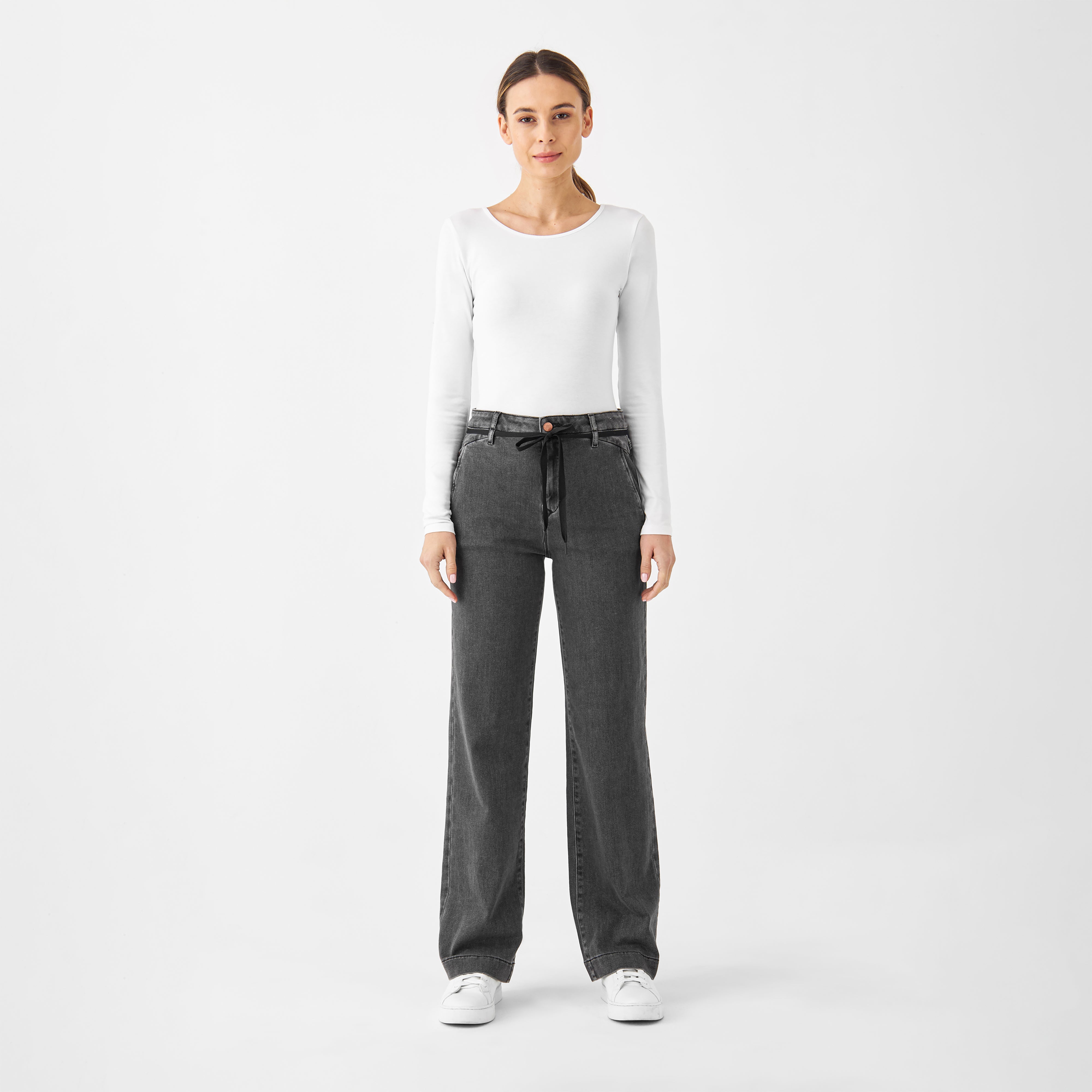 Frontansicht eines Models in weißem Langarmshirt und Sneakern: Medium Grey DEW French Pocket Jeans mit hohem Bund, ausgestelltem Bein und französischen Taschen.