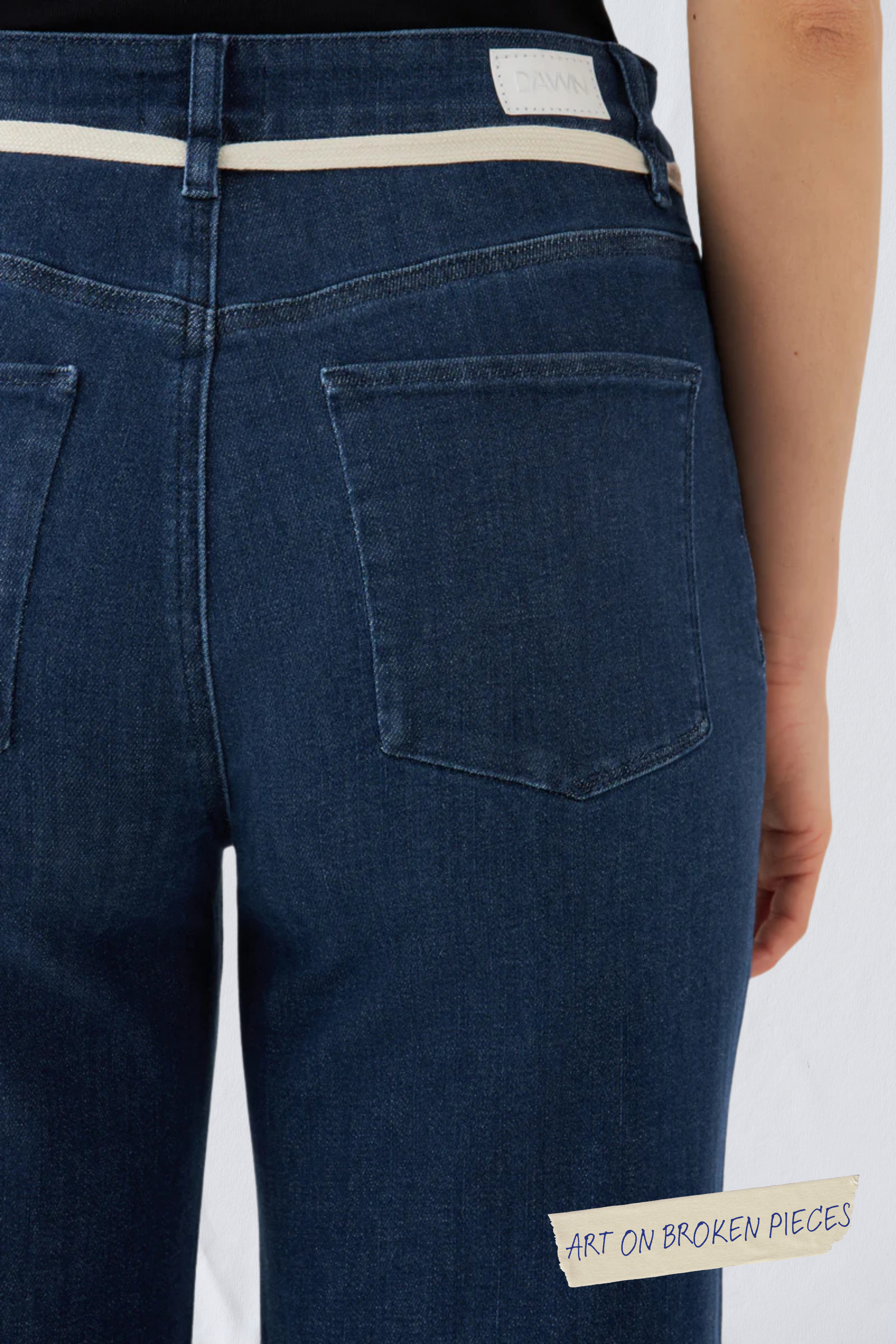 Dunkelblaue, gekürzte DEW Jeans mit hohem Bund und ausgestelltem Bein; Rückansicht mit zwei Gesäßtaschen und DAWN-Label am Taillenband.